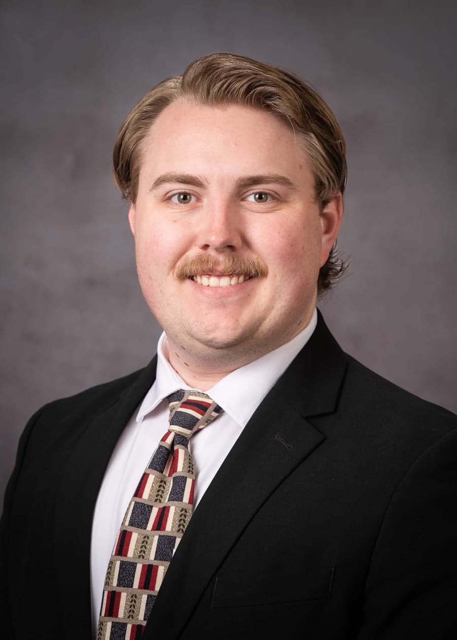 Brigham smiling in a dark suit and a red, navy and cream patterned tie against a gray backdrop