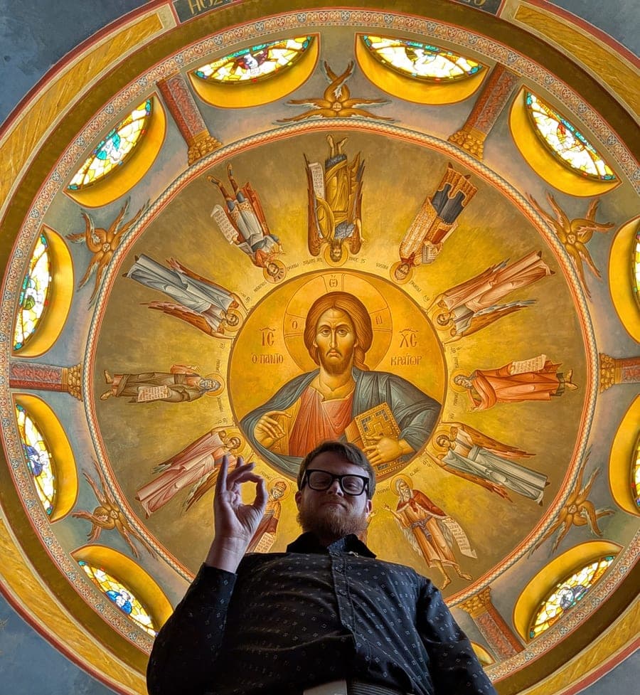 Samuel Baird standing beneath a domed church ceiling painted with Christ Pantocrator, making an OK hand gesture